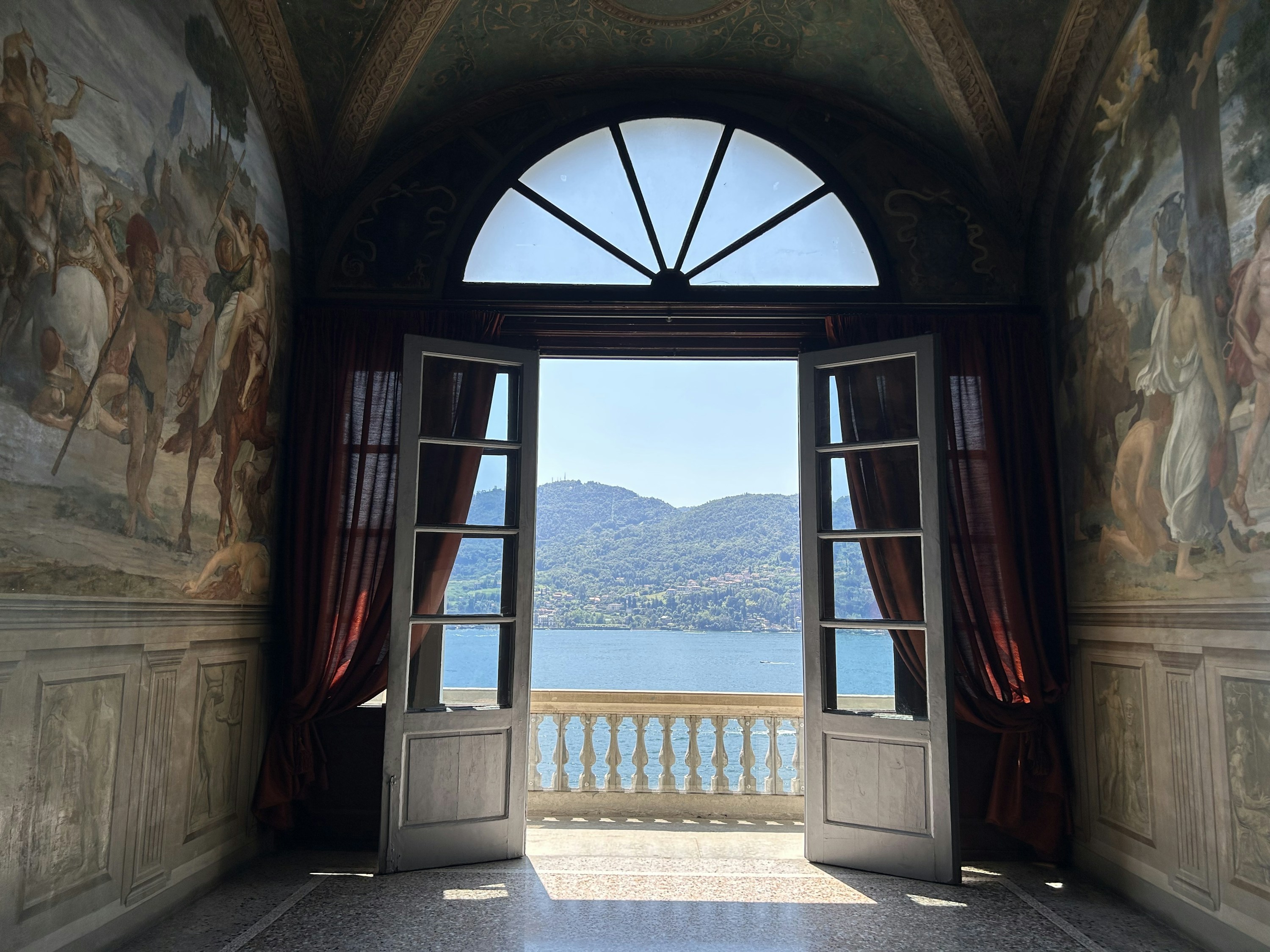 A frescoed room opening through arched doors to an Italian lake